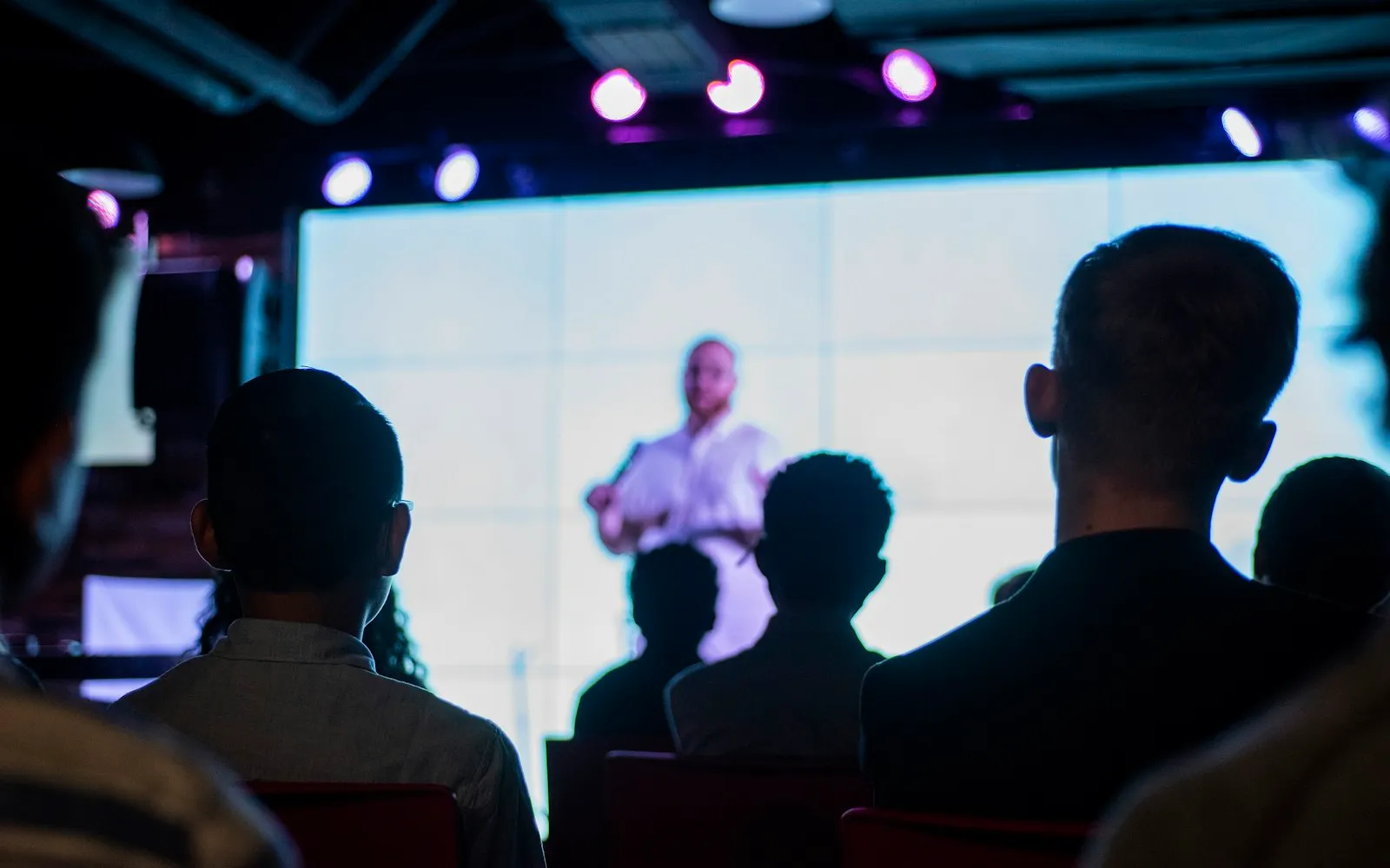 An audience seen from behind watching a speaker on a bright screen in a darkened room with stage lights above