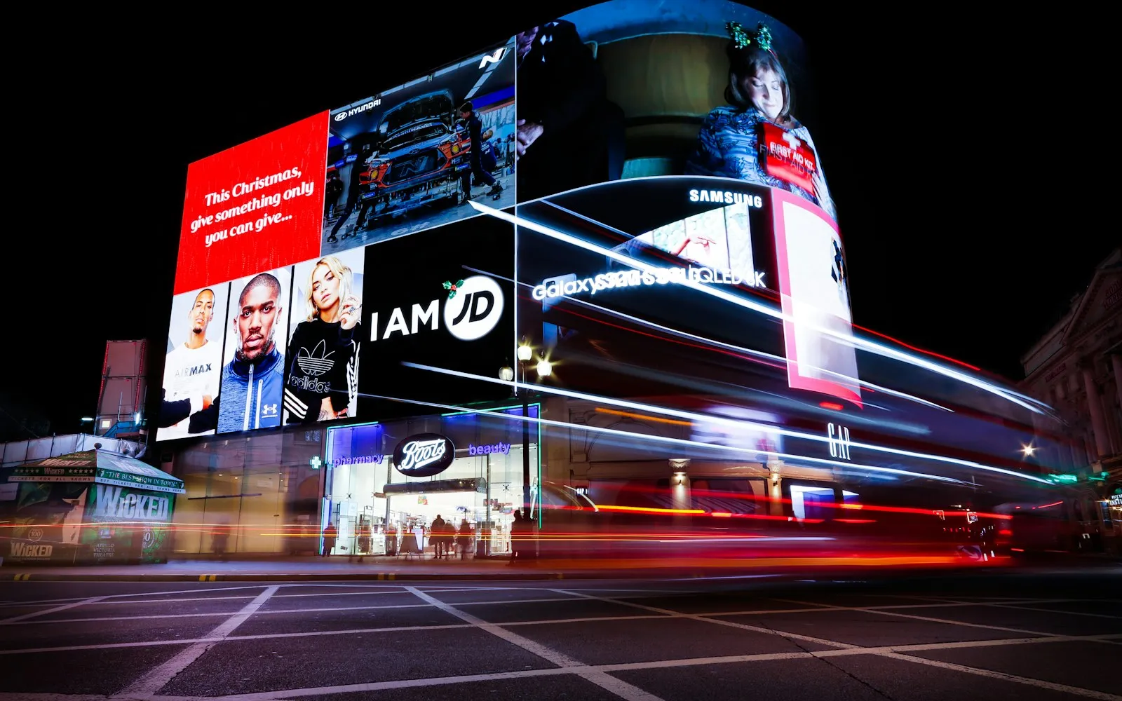A long-exposure night photo of large illuminated digital billboards on a city building, with red light trails from passing traffic