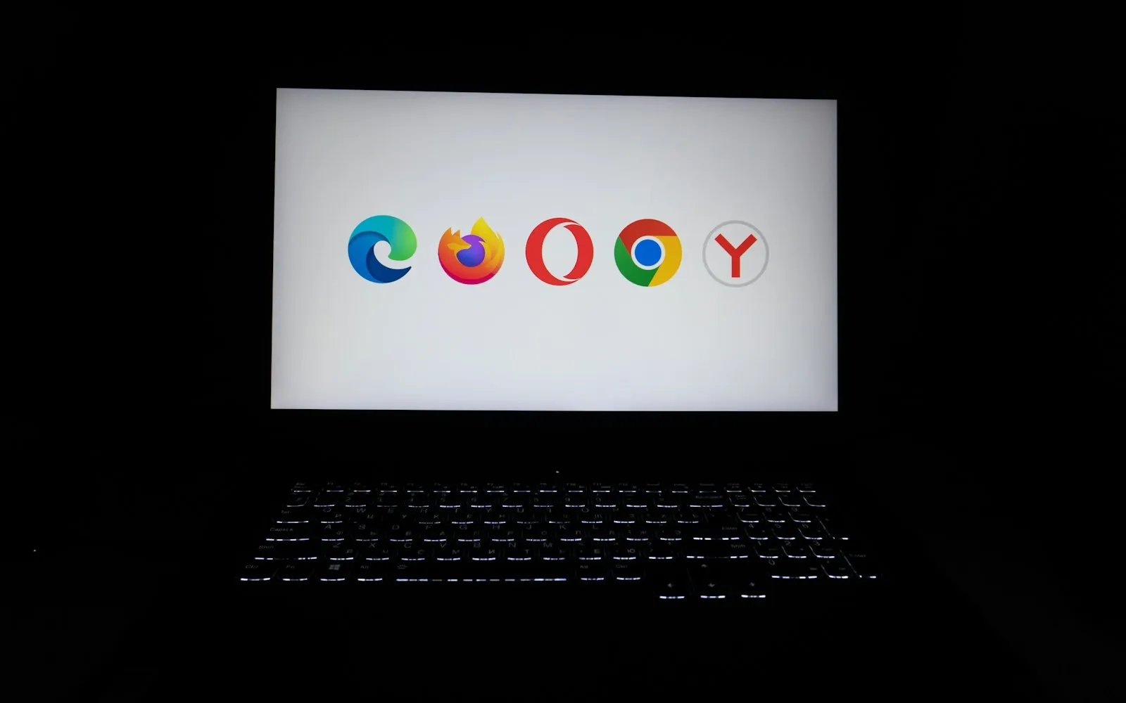 A laptop in a dark room showing the logos of Edge, Firefox, Opera, Chrome and Yandex Browser on a white screen above a backlit keyboard