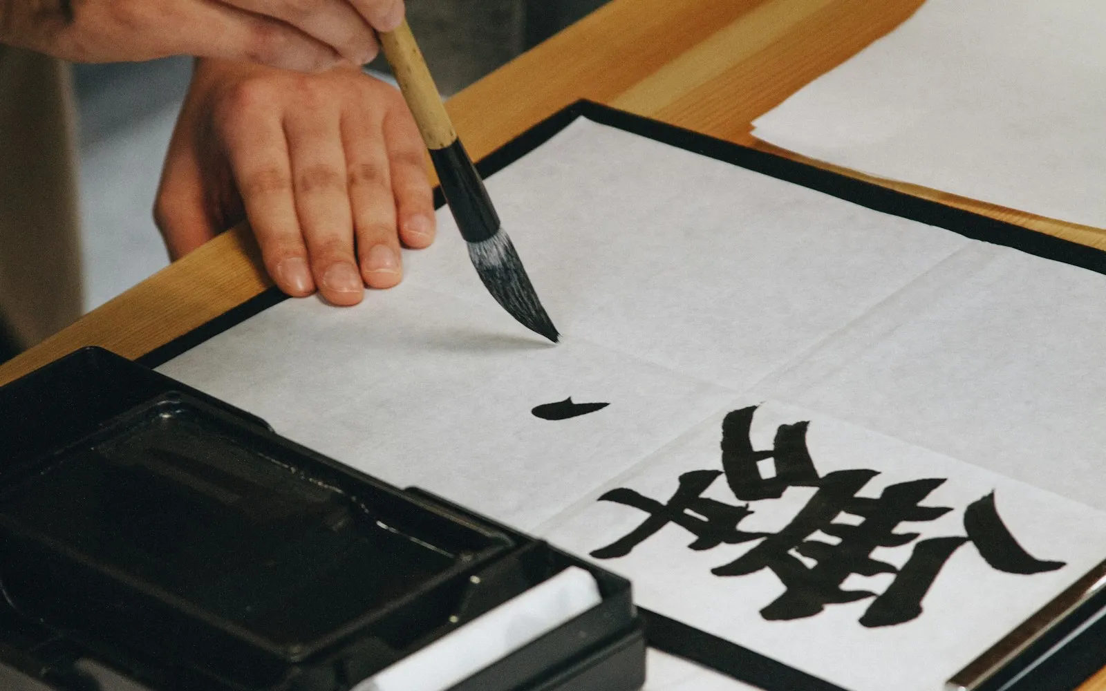 A hand holding a calligraphy brush above a sheet of white paper with a bold black brush-written character beside an ink stone