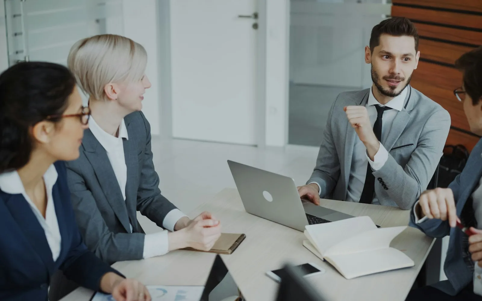 Four colleagues in a bright meeting room talking around a table with a laptop, notebooks and a phone