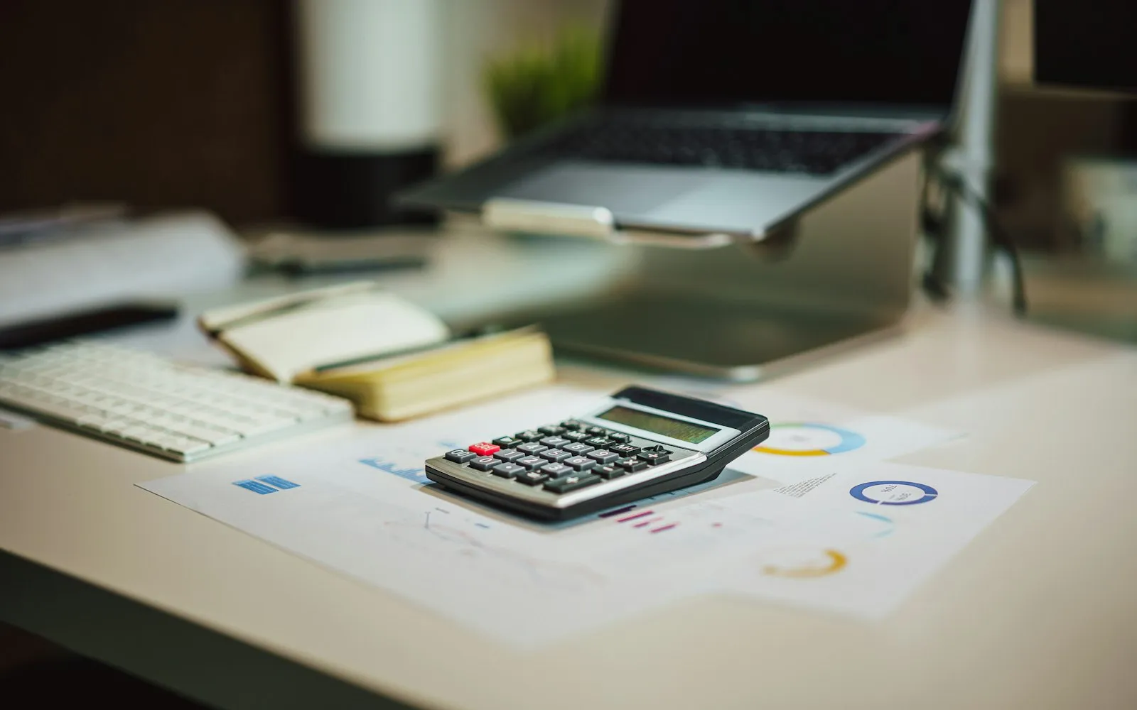 A calculator resting on printed charts on a desk, with a keyboard, a notebook and a laptop behind it