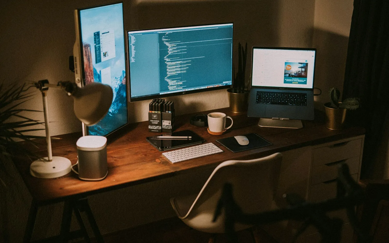 A dimly lit desk with an ultrawide monitor showing code, a laptop on a stand, a keyboard, a lamp and a coffee cup