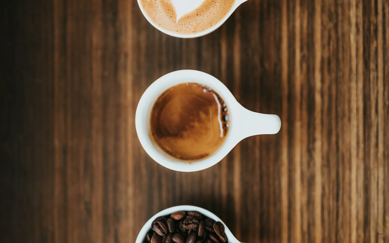 Three white cups seen from above on a wooden table, holding a latte with milk art, an espresso and coffee beans