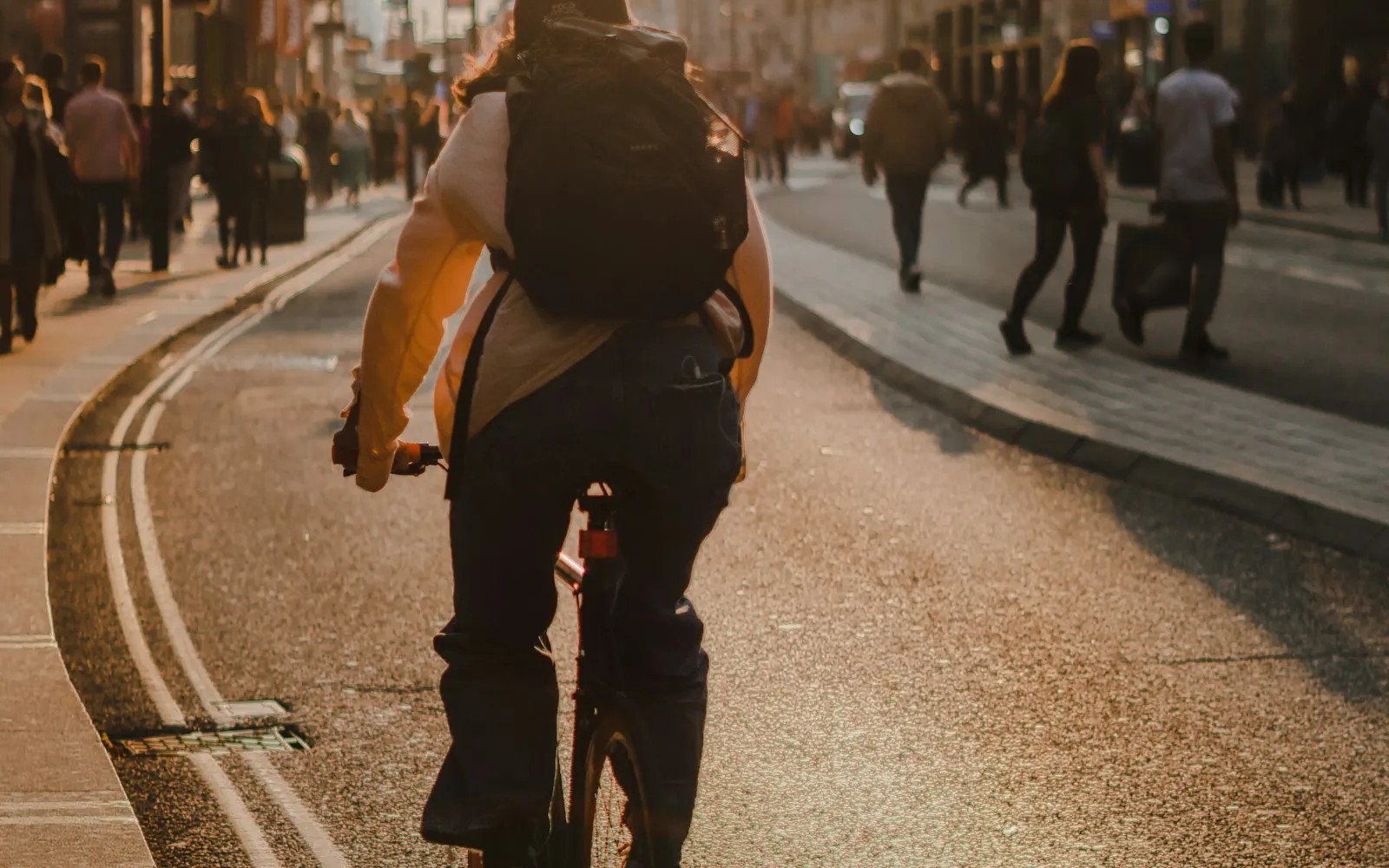 A cyclist with a backpack rides along a busy city street in warm evening light as pedestrians walk by