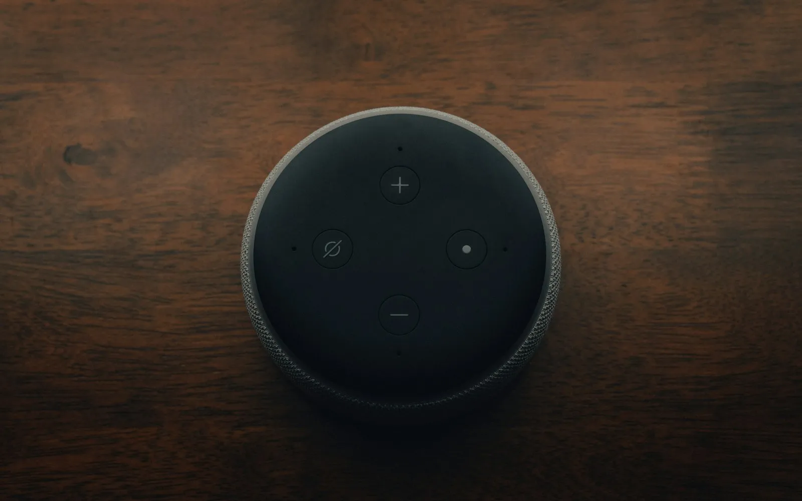 The top of a black Amazon Echo Dot smart speaker with volume, mute and action buttons, seen from above on a dark wooden table