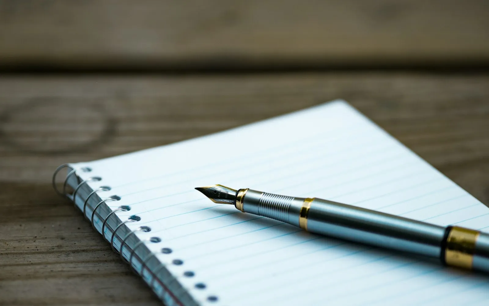 A silver and gold fountain pen lying on a blue-lined spiral notebook on a wooden table