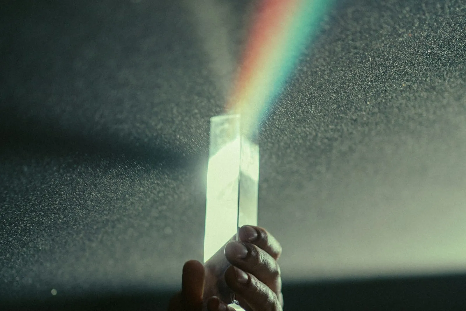 A hand holding up a glass prism that splits light into a rainbow beam