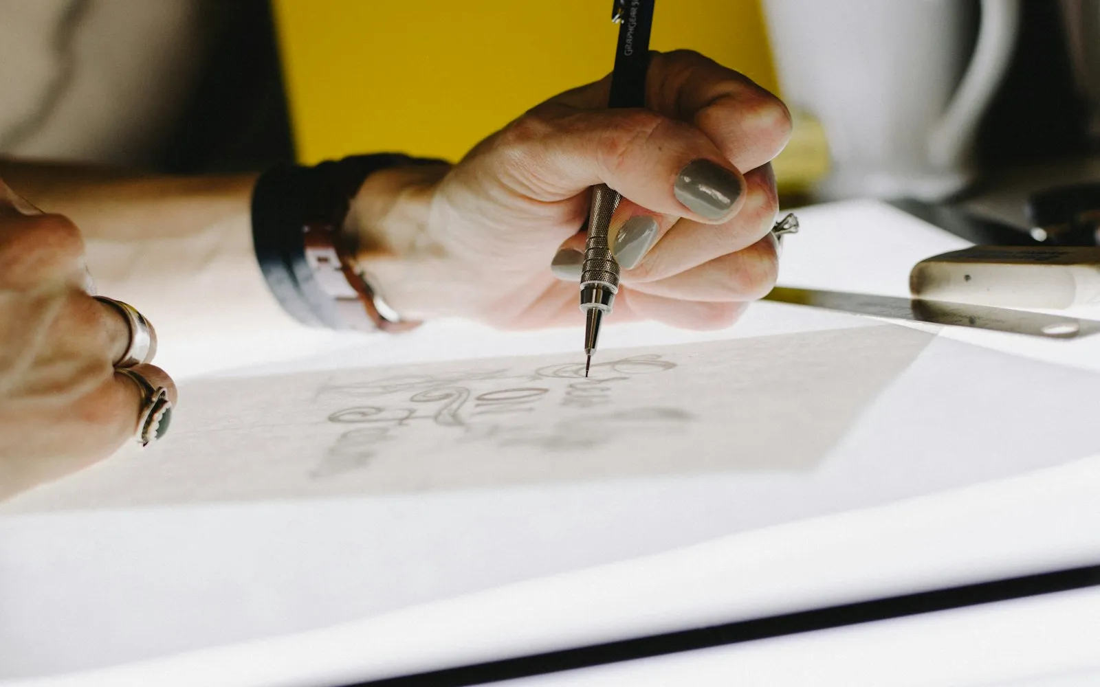 A hand sketching lettering with a mechanical pencil on tracing paper over a lit drawing board