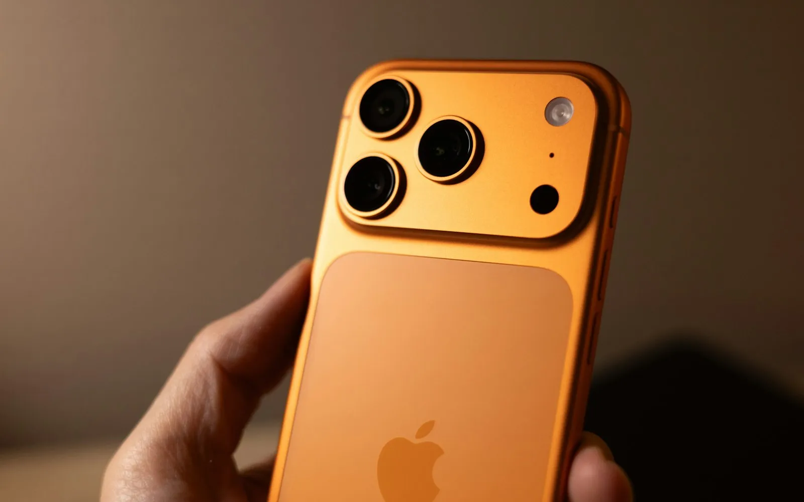 A hand holding an orange iPhone with a triple-lens camera and the Apple logo on the back, against a soft brown background