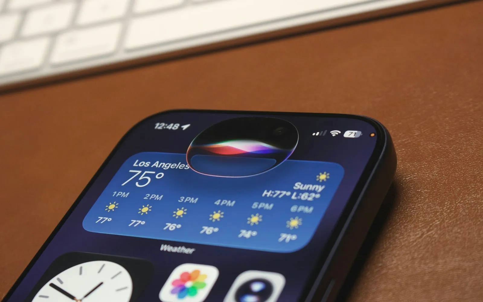An iPhone on a brown leather desk mat beside a keyboard, with the colourful Siri glow at the top of the screen above a weather widget