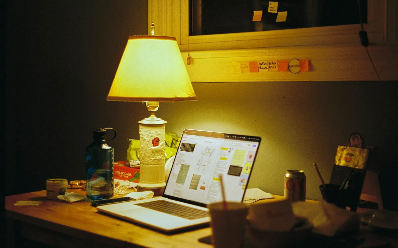 A laptop open on a wooden desk lit by a yellow table lamp at night