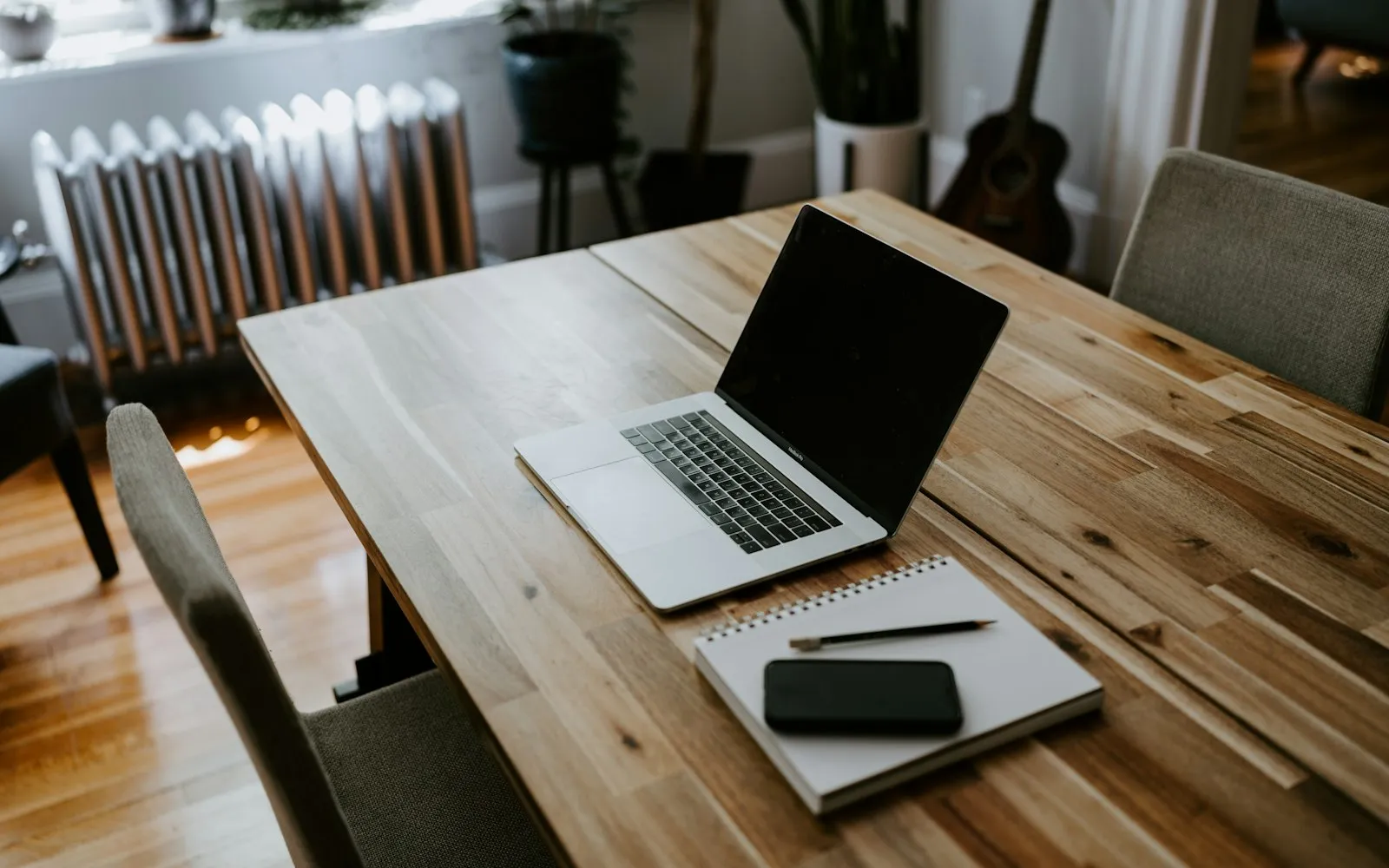An open laptop, a spiral notebook with a pencil and a phone on a wooden table in a living room