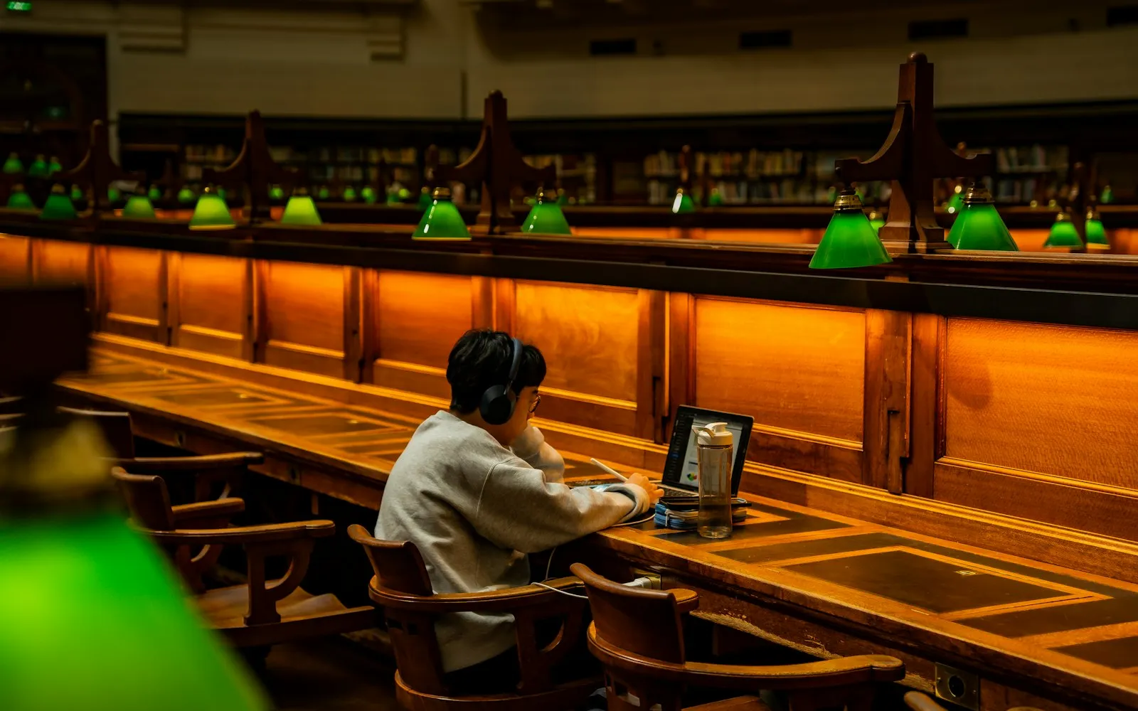 A person working on a laptop at a long wooden desk with green reading lamps in a large library