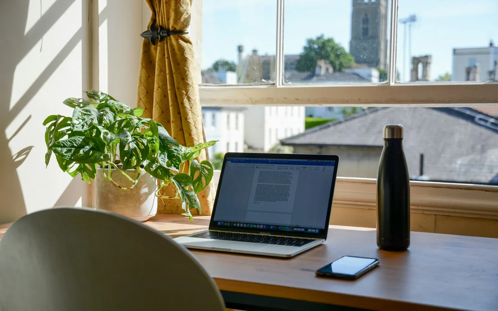 A laptop open to a document on a wooden desk by a sunlit window, next to a potted plant and a water bottle