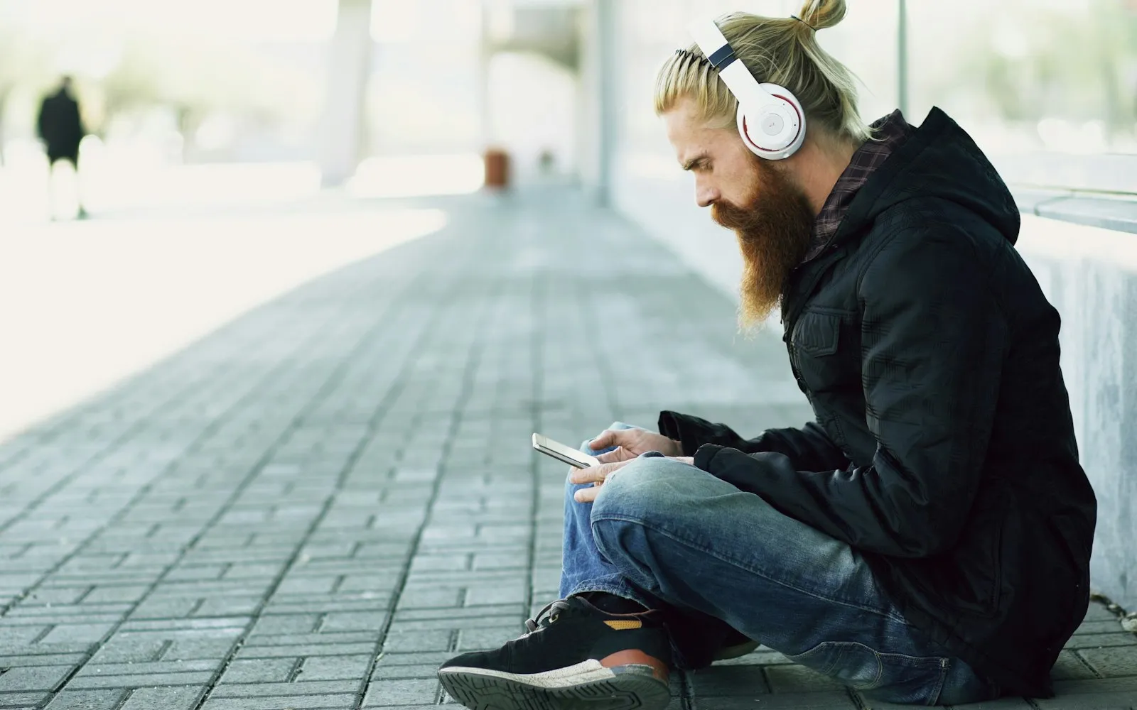 A bearded man in a black coat sitting on a pavement wearing white headphones and looking at a smartphone