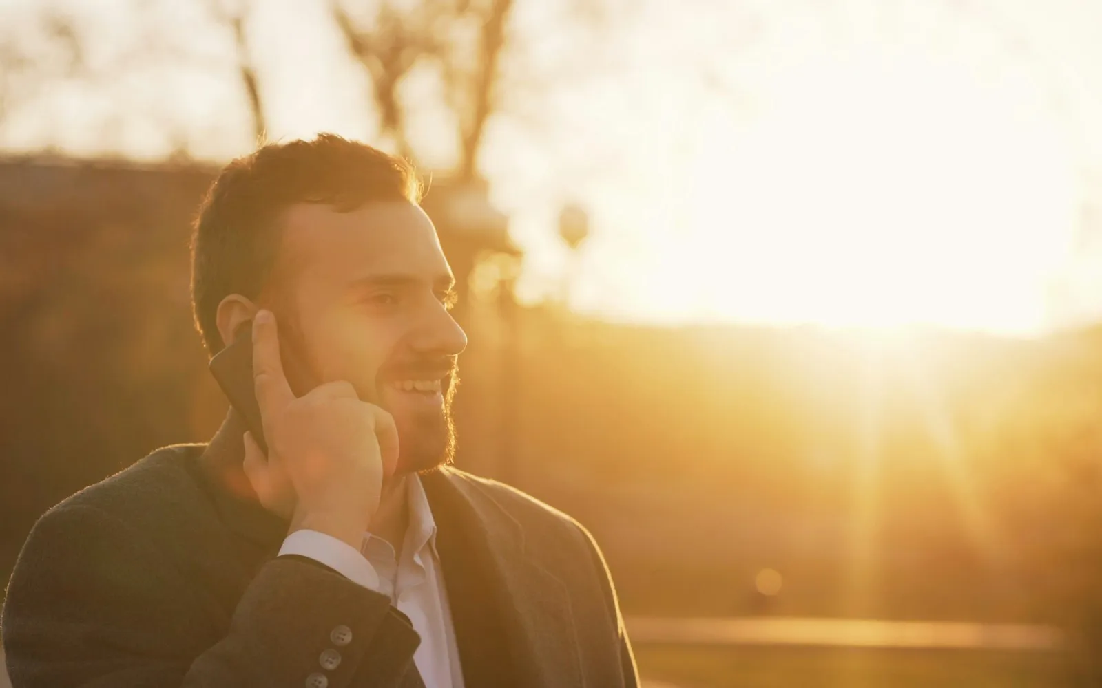 A smiling man in a jacket holding a phone to his ear outdoors, backlit by a low sun
