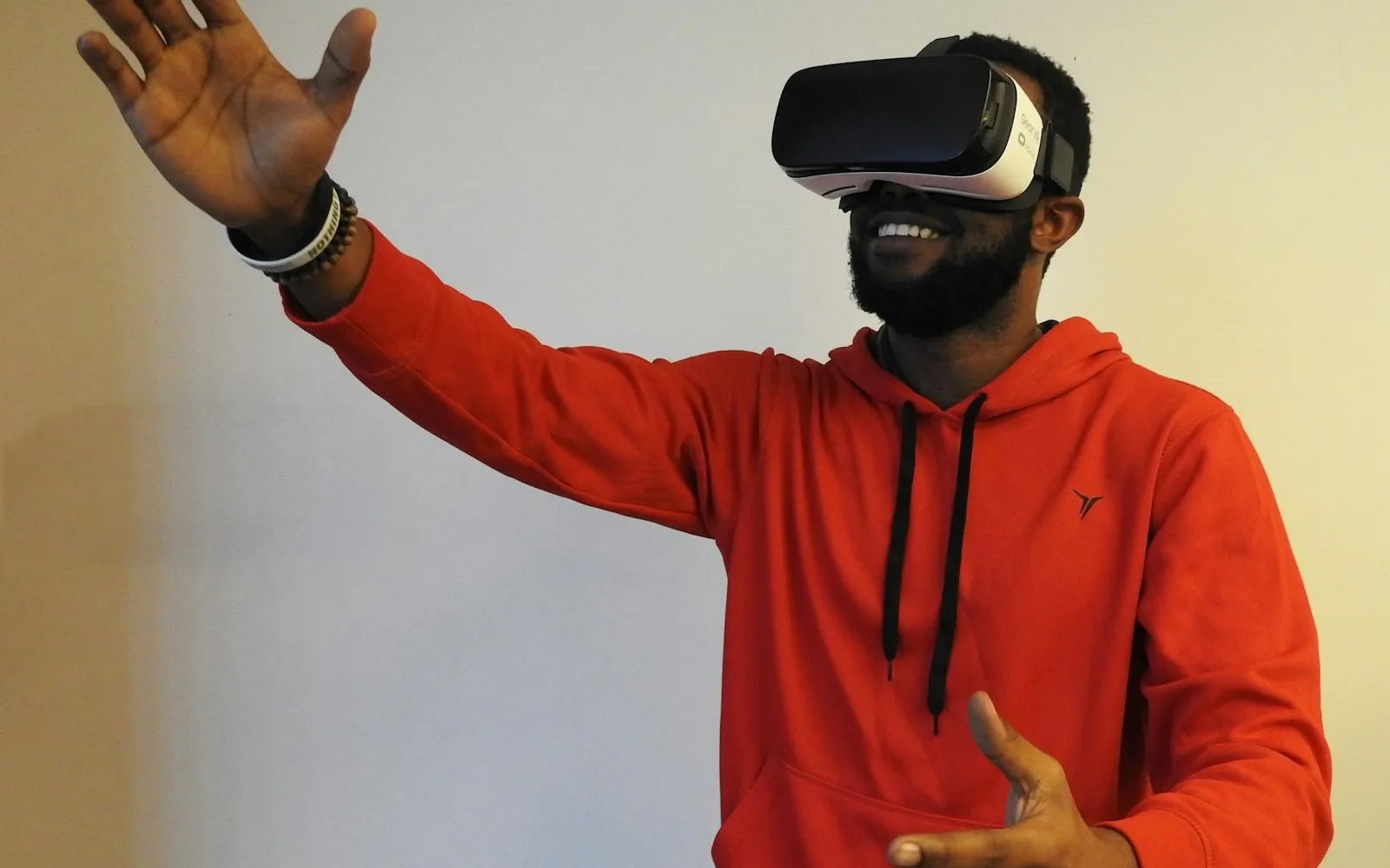 A smiling man in a red hoodie reaches out one hand while wearing a virtual reality headset