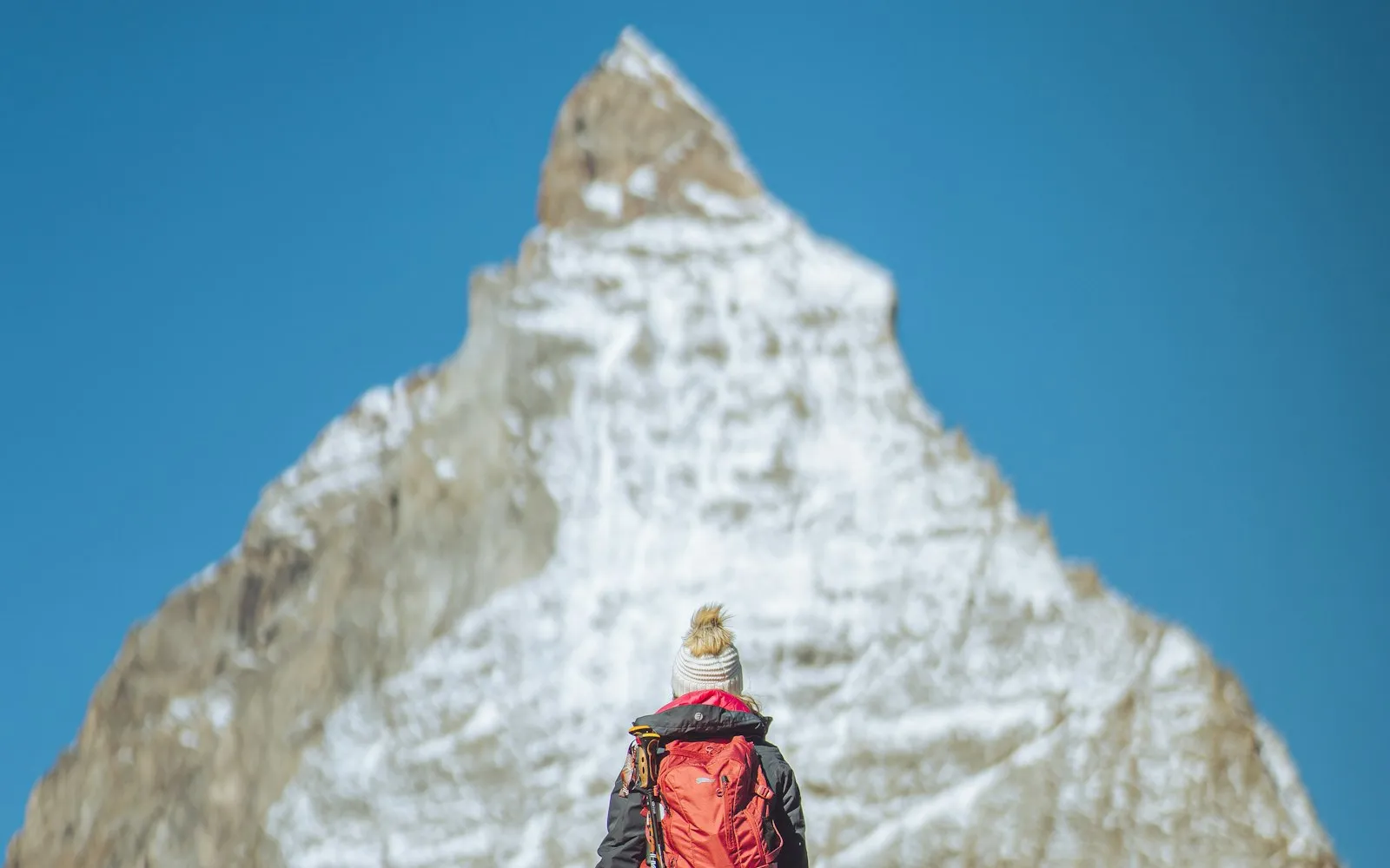 A hiker in a red jacket and backpack, wearing a white bobble hat, looks up at a snow-dusted mountain peak against a clear blue sky
