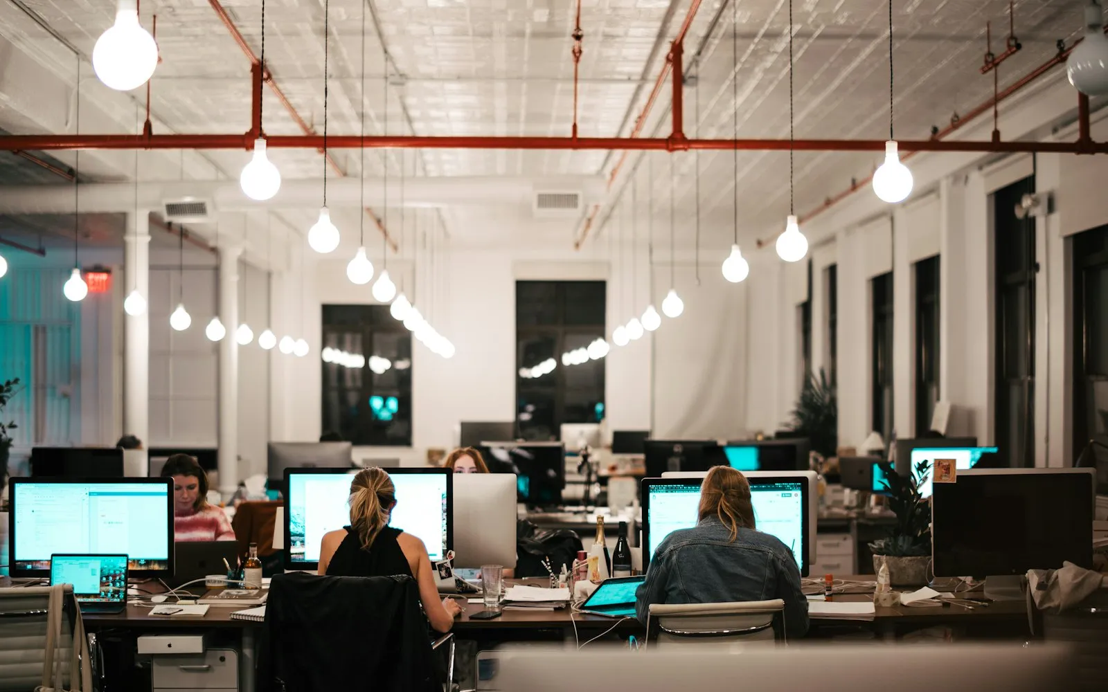 An open-plan office with people working at rows of computer monitors under hanging light bulbs