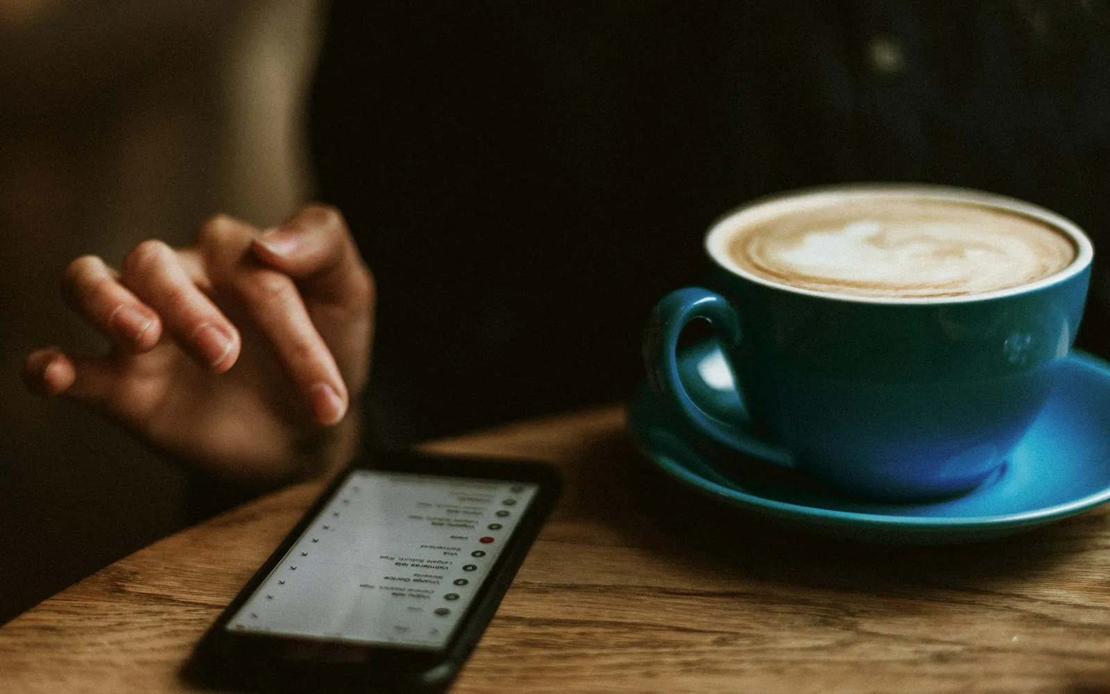 A hand gesturing over a smartphone on a wooden table next to a blue cup of latte