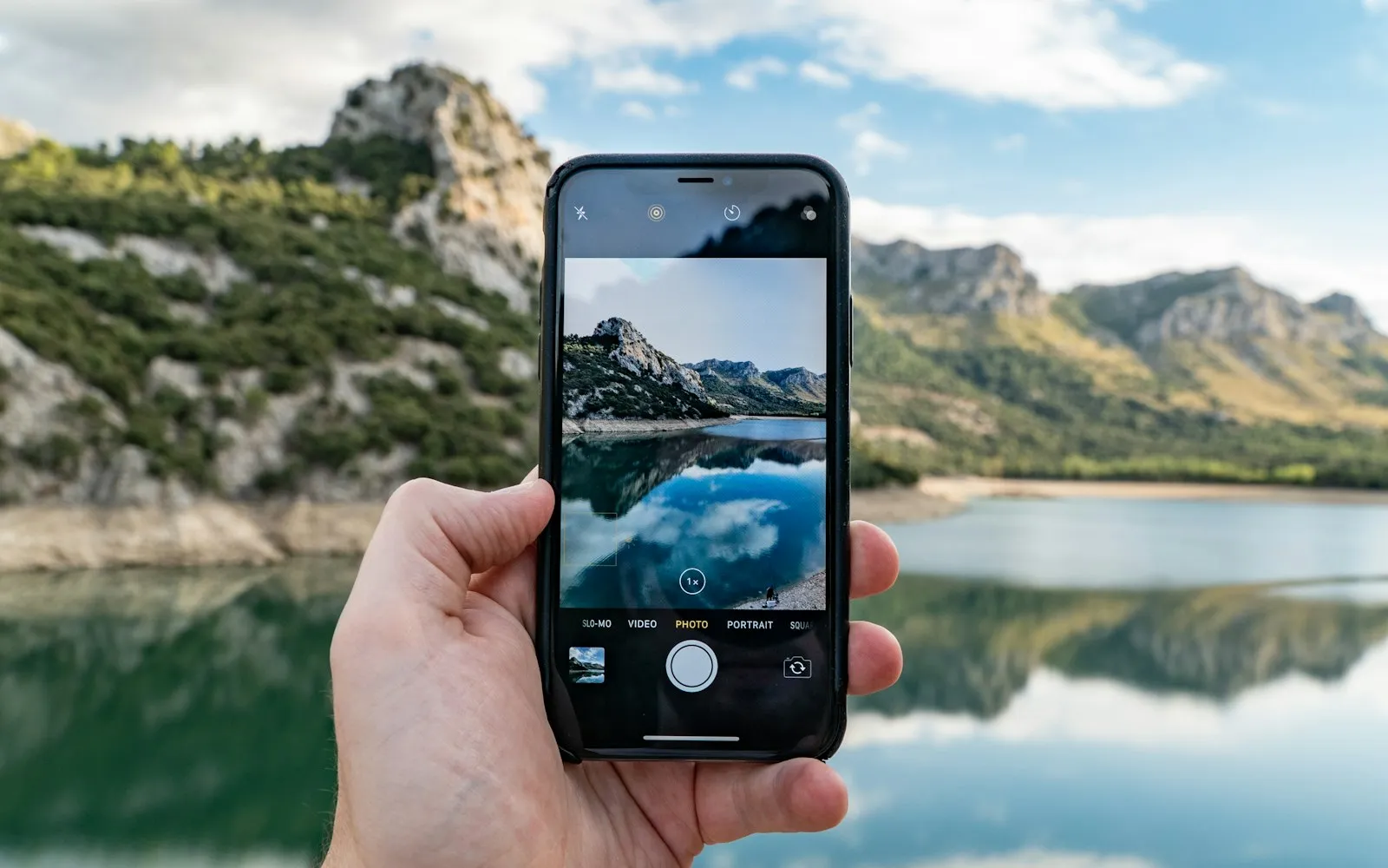 A hand holding a smartphone up to photograph a mountain lake, with the scene shown on the phone's screen