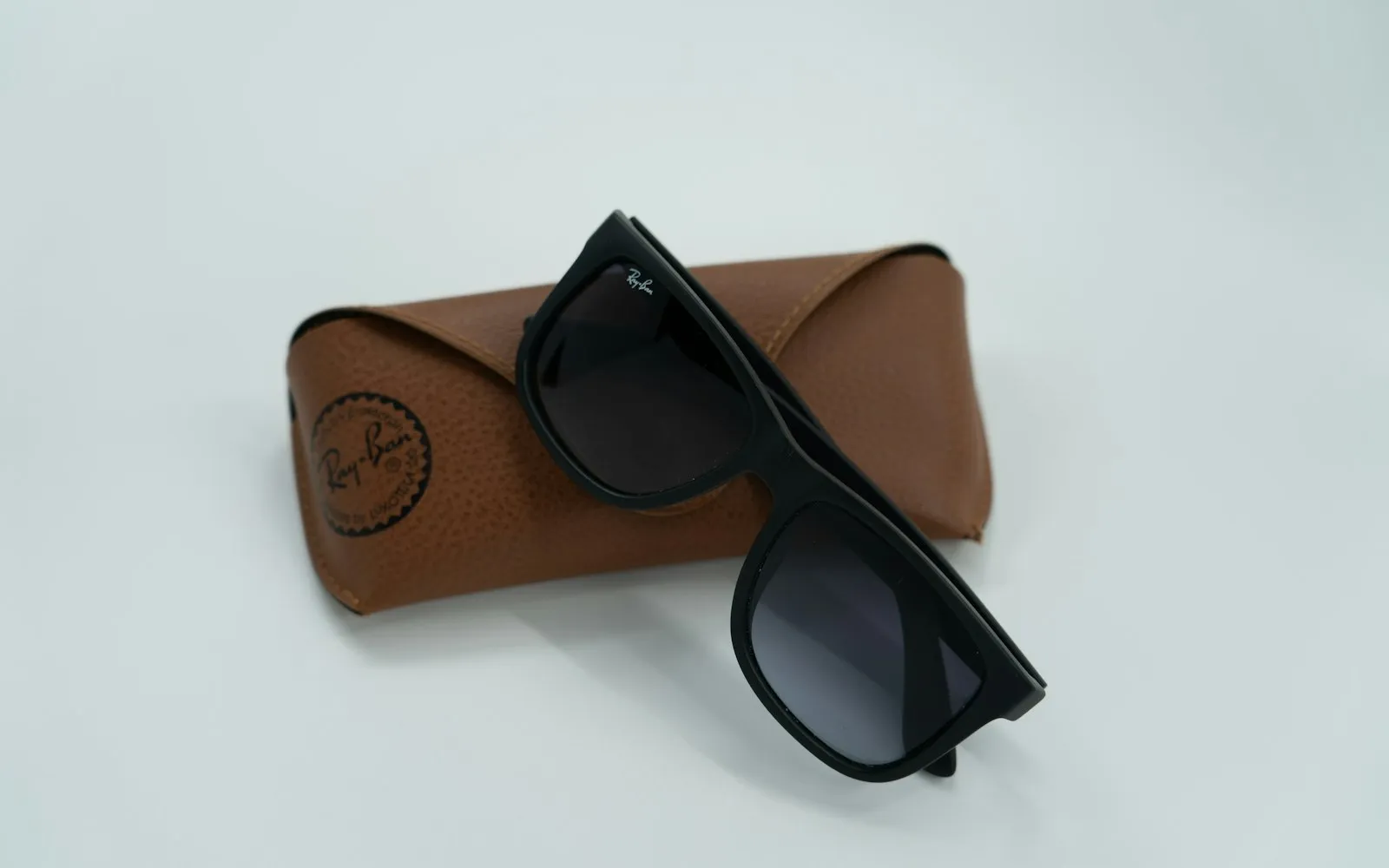 A pair of black Ray-Ban sunglasses resting on a brown leather Ray-Ban case against a pale background