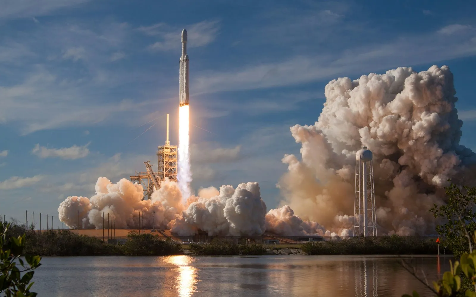 A tall rocket lifting off from a launch pad in a plume of white smoke under a blue sky with a water tower to the right