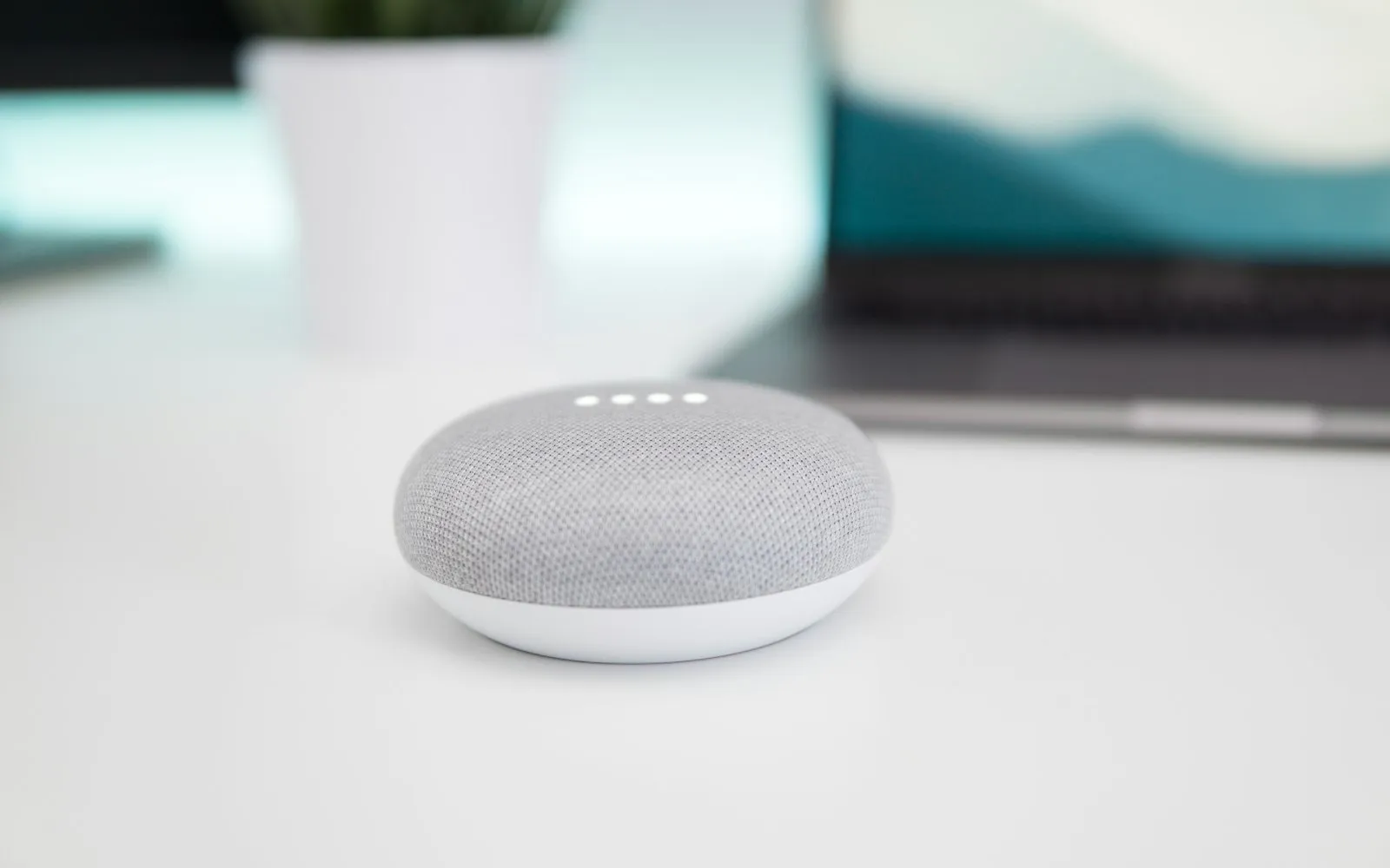 A small grey fabric smart speaker with four lit indicator dots on a white desk, with a laptop out of focus behind it