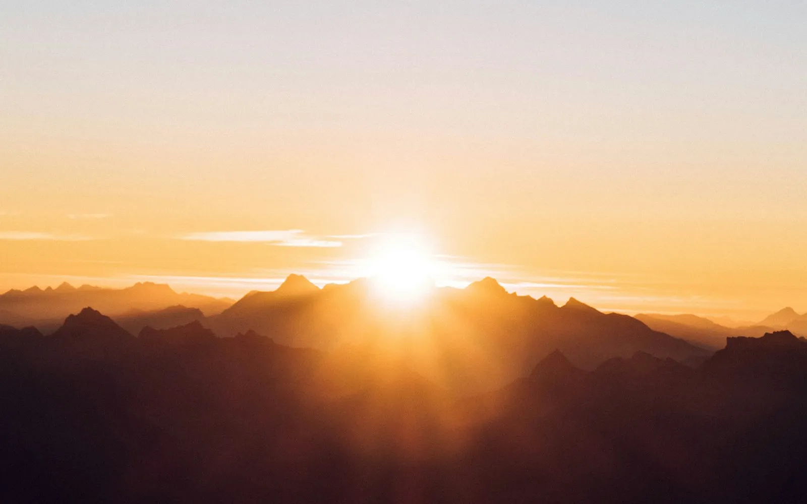 The sun low on the horizon glowing behind layered mountain ridges under a pale orange sky