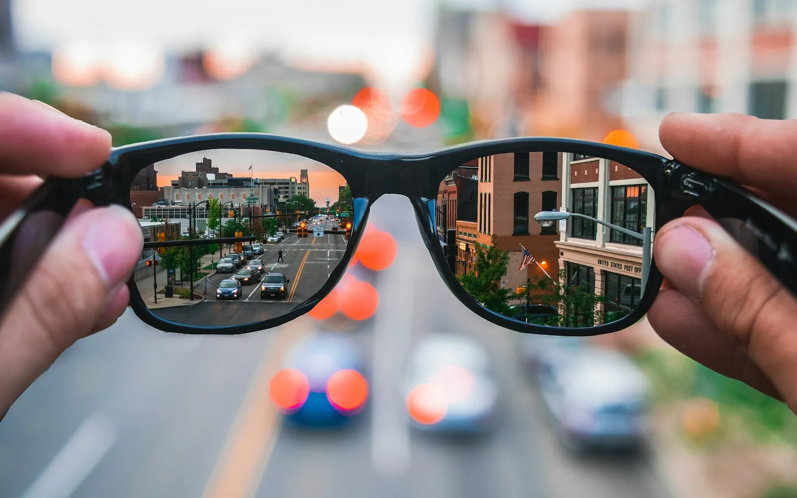 A pair of black-framed glasses held up to a city street at dusk, with the road and buildings in focus through the lenses
