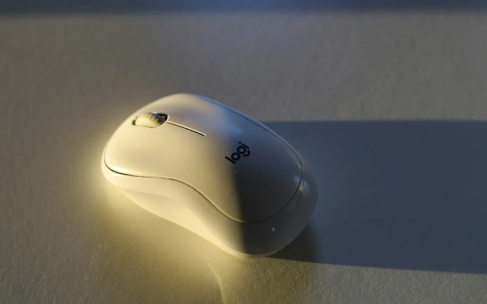 A white wireless computer mouse on a pale desk lit by warm light and casting a long shadow