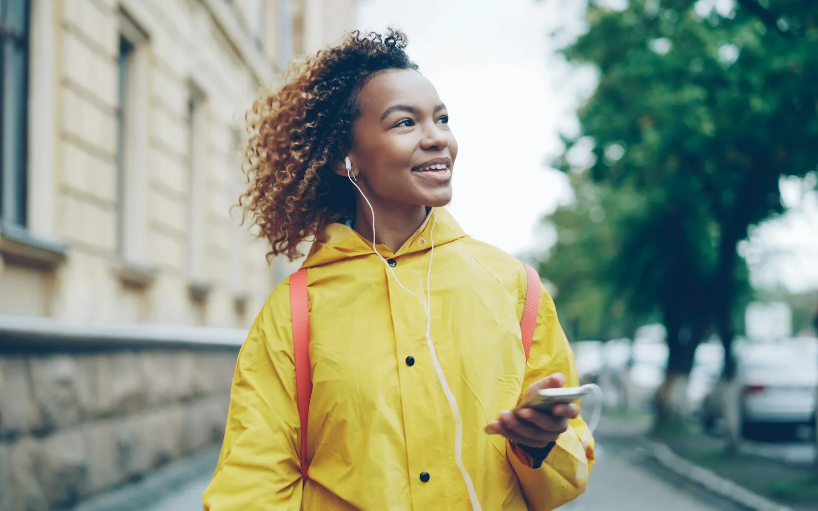 A smiling woman in a yellow raincoat wearing earphones and holding a smartphone as she walks down a city street