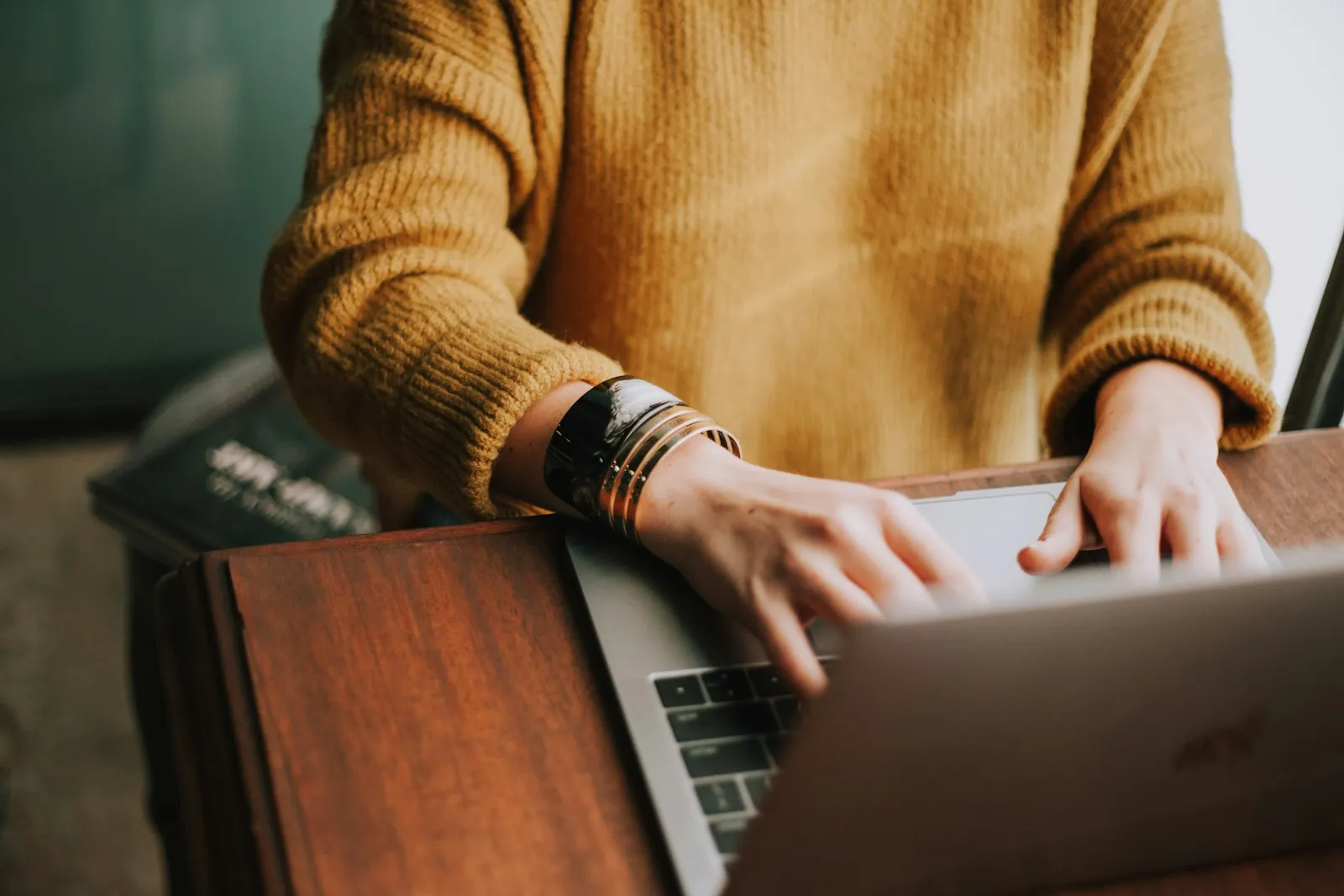 A person in a mustard sweater typing on a laptop at a wooden table